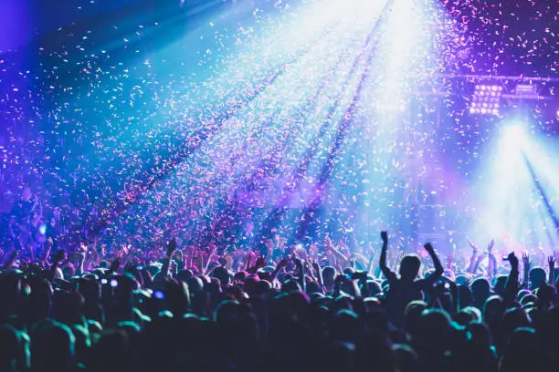 Photo of A crowded concert hall with scene stage lights, rock show performance, with people silhouette, colourful confetti explosion fired on dance floor air during a concert festival