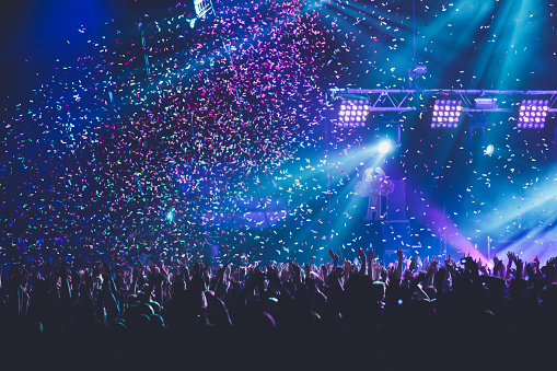 A crowded concert hall with scene stage lights, rock show performance, with people silhouette, colourful confetti explosion fired on dance floor during a concert festival
