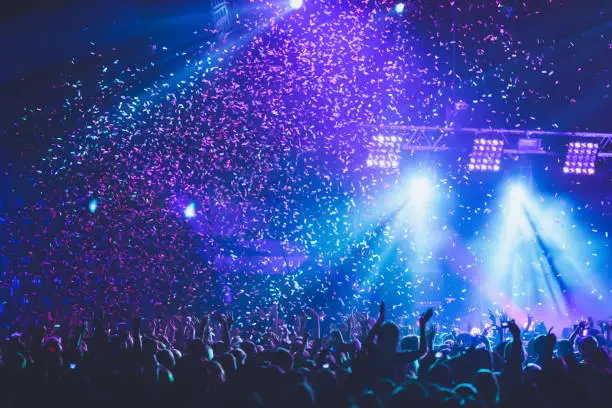 Photo of A crowded concert hall with scene stage lights, rock show performance, with people silhouette, colourful confetti explosion fired on dance floor air during a concert festival