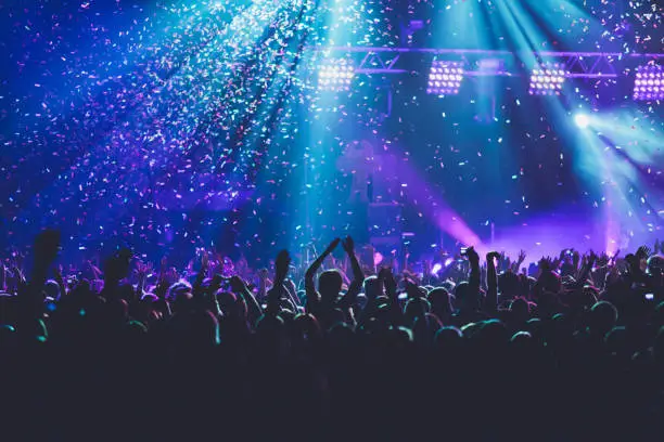 Photo of A crowded concert hall with scene stage lights, rock show performance, with people silhouette, colourful confetti explosion fired on dance floor air during a concert festival
