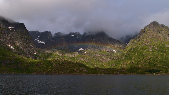 Beautiful view of strong colorful rainbow on the shore of Raftsundeet strait in front of Austvågøya island, Lofoten, Norway with majestic rugged mountains covered by green plants on cloudy summer day.