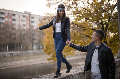 Loving young couple in leather jacket and bandana on forehead enjoying time together in the city on sunny autumn day, holding hands while she walking across stone wall