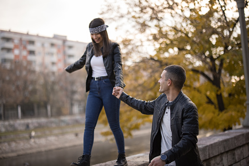 Loving young couple in leather jacket and bandana on forehead enjoying time together in the city on sunny autumn day, holding hands while she walking across stone wall