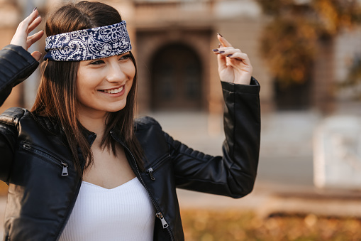 One young woman in leather jacket and bandana on forehead walking in the city on sunny day