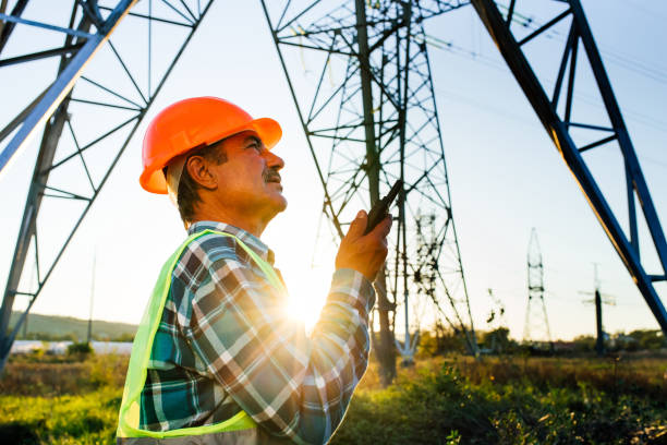 istock Low angle side view of middle aged male technician in hardhat speaking on walkie talkie while standing in back lit against electric power poles 1289542812