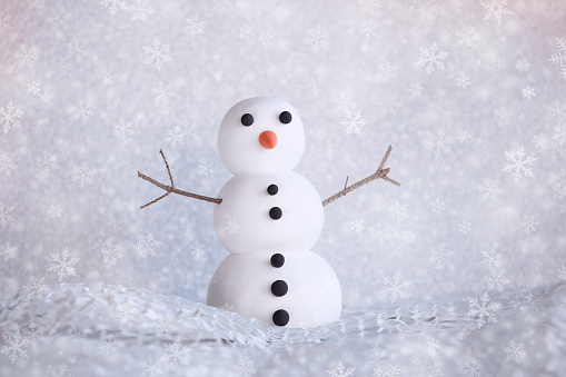 Funny small snowman figurine with carrot, brunches and black buttons, puppet in winter background. Snow falling down.