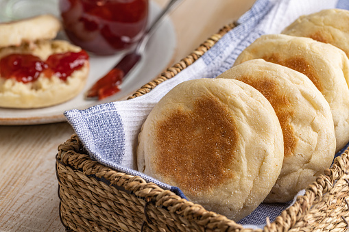 English muffins in a basket with a muffin and preserves in background on a wooden table
