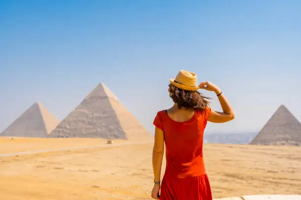 Photo of A young tourist in a red dress looking at the Pyramids of Giza, the oldest Funerary monument in the world. In the city of Cairo, Egypt