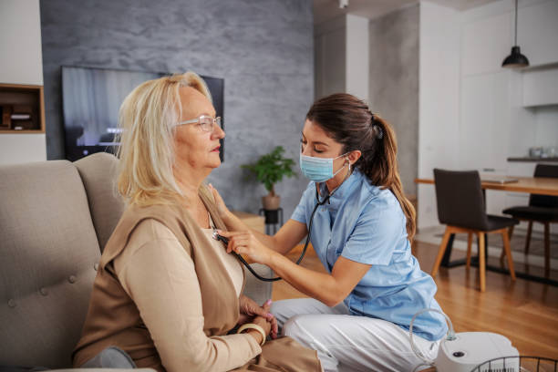 istock Nurse with protective mask on sitting next to senior woman and using stethoscope to check on lungs during corona outbreak. 1289565697