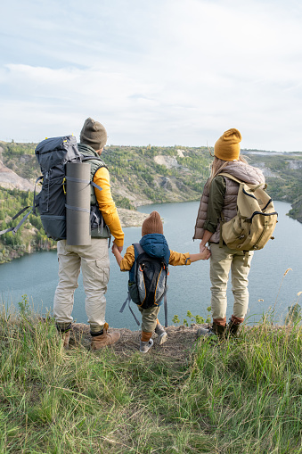 Rear view of young couple and their little son with backpacks holding by hands while standing on grass and looking at lake between mountains