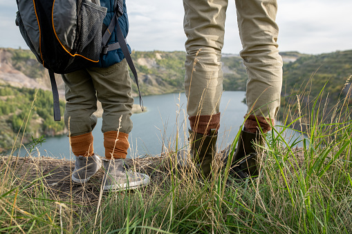 Rear view of legs of young mother and little son with backpacks standing on the ground in front of lake or river surrounded by mountains