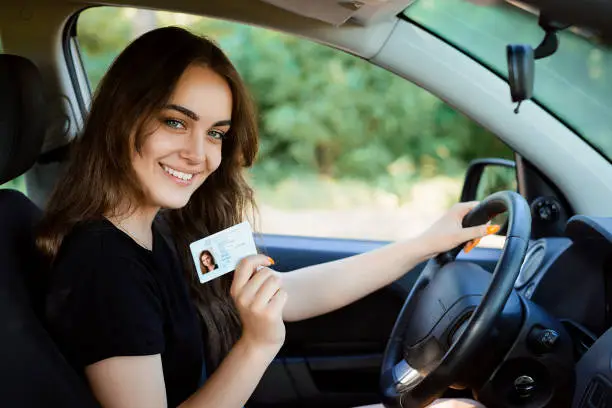 Photo of Smiling young female with pleasant appearance shows proudly her drivers license, sits in new car, being young inexperienced driver, looks with joyful expression