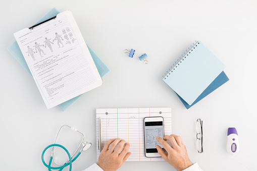 Overview of medical worker hands on blank card and touchscreen of gadget while scrolling through data in smartphone by workplace