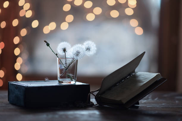 Three dandelions in a glass and a book on a wooden table, a dark background, close up.