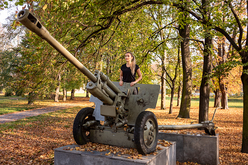 Young attractive blond woman standing on the carriage of a historic World War II cannon and looking ahead. It is a Russian cannon that stands on a concrete pedestal in the park.