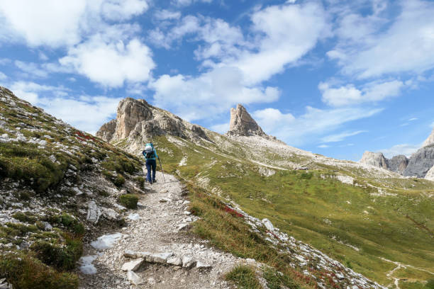 istock A woman with backpack and sticks hiking on a narrow path in Italian Dolomites. There are sharp and steep mountains. At the bottom of a small valley there is a small, navy blue lake. Raw landscape. 1289575798