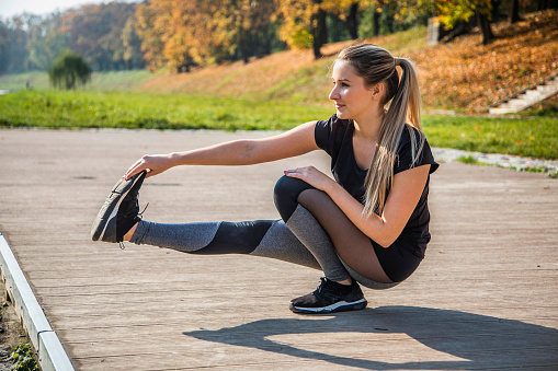 Young attractive blond woman stretching in the city pier on the river. She squatting on her left leg and her right one is pulled in the air in front of her. She wipes it by the tip of his foot. She looking ahead, has wearing a black T-shirt and grey sport leggings. Autumn sunny day.