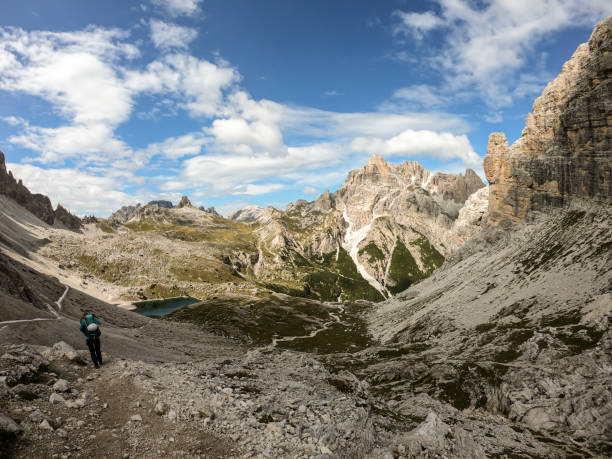 istock A woman with backpack and sticks hiking on a narrow path in Italian Dolomites. There are sharp and steep mountains. At the bottom of a small valley there is a small, navy blue lake. Raw landscape 1289577002