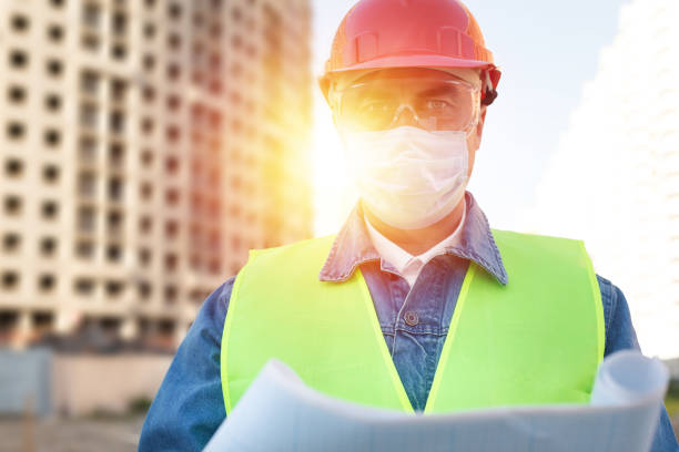 istock Engineer in red protective helmet and medical mask with blueprint  at construction site 1289578207