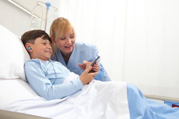 il bambino felice sdraiato a letto nella stanza d'ospedale e l'infermiera sorridente che naviga su internet indossando auricolari - child text messaging little boys male foto e immagini stock