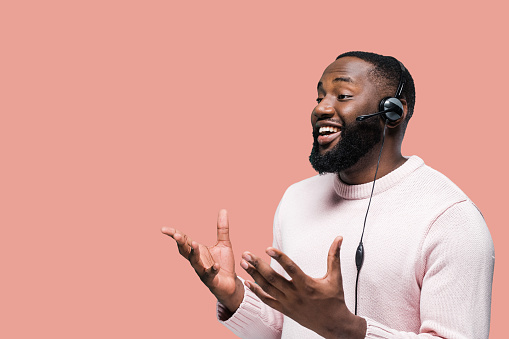 Trabajador afroamericano del centro de llamadas con auriculares en ropa casual de pie sobre un fondo rosado aislado mientras se comunica con un cliente y sonríe photo
