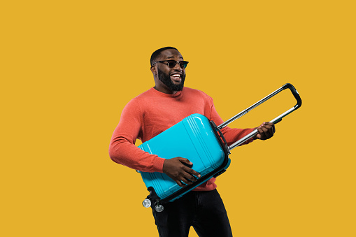 Cheerful African American guy in a sunglasses and in casual wear holds a blue suitcase in his hands standing on isolated orange background. In anticipation of travel