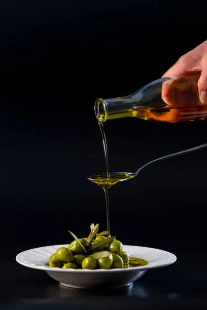 Photo of view of pure olive oil pouring on a metal spoon and overflowing into a plate with tempting olives