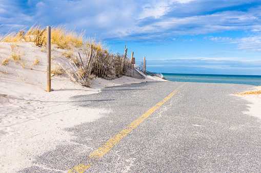 A sand covered paved road with a yellow center line passes a grass covered dune and sand fence as it heads to the sea beyond in Dennis, Massachusetts on an early December morning.