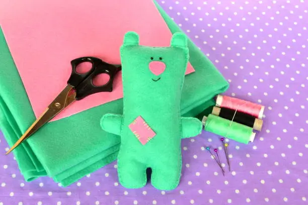 Photo of Cute green felt Teddy bear, handmade children toy diy. Scissors, color rolls threads, needles - sewing kit, tailor workplace table. Smiling soft baby teddy bear toy photo. Kids handicrafts background