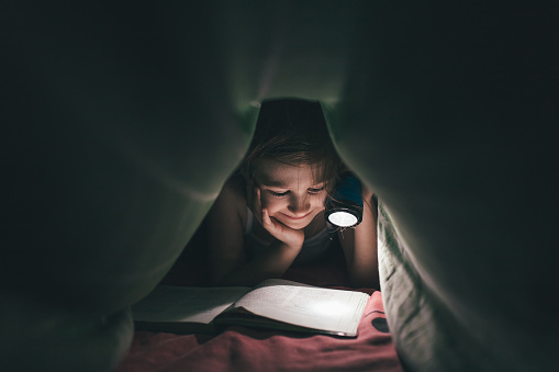 smile little girl is read a book under a blanket with a flashlight in a dark room at night,  in bed