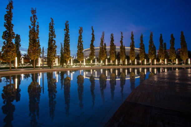 Blue Hour -view on  the park "Krasnodar", Krasnodar.  A row of cypresses and a reflection in a shallow pool. Blue Hour -view on  the park "Krasnodar", Krasnodar.  A row of cypresses and a reflection in a shallow pool. Soccer Stadium at far distance. Illuminated park and stadium krasnodar stock pictures, royalty-free photos & images