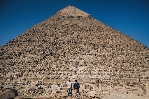 Two men stand in front of the old pyramid with their pants down.