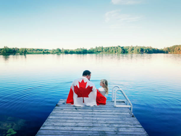 vater und tochter in große kanadische flagge gewickelt sitzen auf holzdock am see. kanada-tag-feier im freien. vater und kind sitzen am 1. juli zusammen, um den nationalen kanadatag zu feiern. - canada day fotos stock-fotos und bilder