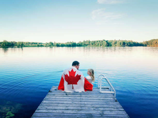 padre e hija envueltos en una gran bandera canadiense sentado en un muelle de madera junto al lago. celebración del día de canadá al aire libre. papá y niño sentados juntos el 1 de julio celebrando el día nacional de canadá. - canada canada day canadian flag canadian culture fotografías e imágenes de stock