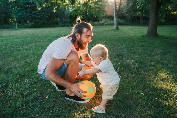 Photo of Fathers Day. Father playing ball with toddler baby boy outdoor. Parent spending time together with child son in a park. Authentic lifestyle tender moment. Happy dad and active family life.