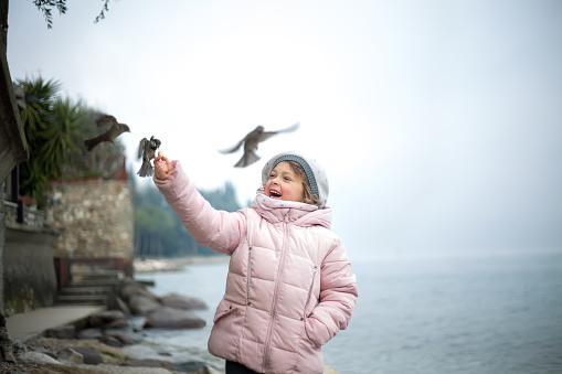 A little girl hand-feeds the sparrows on the shores of Lake Garda. Happy child outdoors