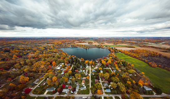Beautiful full panoramic aerial view of the heart shaped Saugany Lake in Indiana surrounded by residential homes and autumn colored trees or foliage with fluffy white clouds in the sky above.