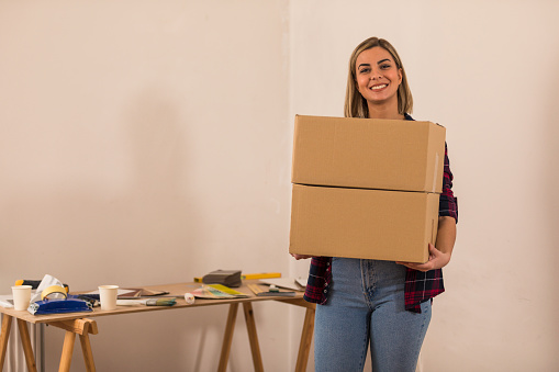 Copy space shot of joyful young woman smiling at camera while holding boxes and moving into her new home.