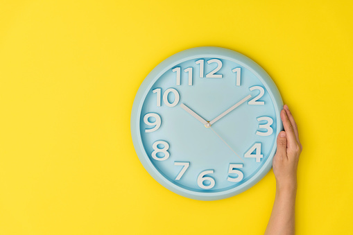 Human hand holding a clock on yellow background.
