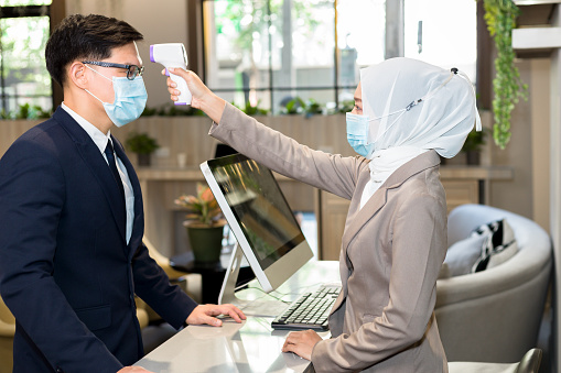 Muslim female receptionist measures tourists' temperatures before staying in a hotel.
