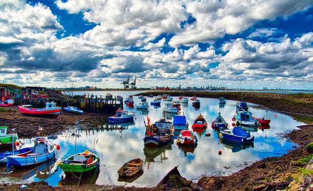 Small fishing boats at anchor in little harbour with dramatic sky, Paddy' Hole, South Gare, Teeside, England, Britain Peaceful small harbor with still water and reflections with cranes of Teesport in background teesside northeast england stock pictures, royalty-free photos & images