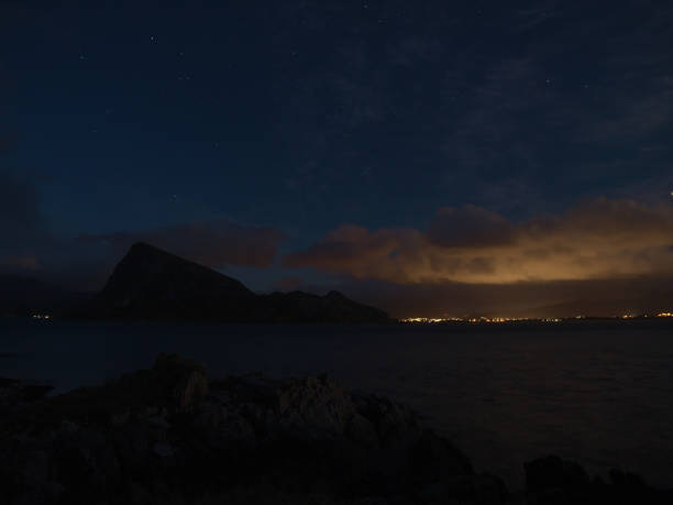 bela vista noturna da costa oeste da ilha de vestvågøy com estrelas no céu, as silhuetas de montanhas e nuvens iluminadas pelas luzes da cidade de leknes em lofoten. - vestvagoy - fotografias e filmes do acervo