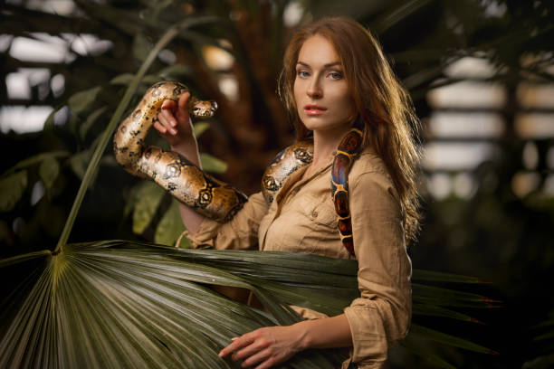 A beautiful girl with natural make-up and red hair stands in the jungle among exotic plants with snake Attractive caucasian woman with python. Beautiful ginger girl with snake in tropical garden. Attractive Caucasian woman with long wavy red hair wearing in khaki. Freedom, harmony with the nature. Beauty girl outdoor. Natural concept Boa stock pictures, royalty-free photos & images