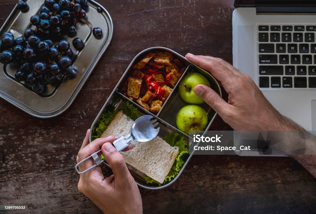 Close up of man hands at desk with a laptop at home or in office with healthy vegan meal in reusable stainless steel lunch box. Healthy lifestyle, zero waste and sustainable plastic free lifestyle Close up of man hands at desk with a laptop at home or in office with healthy vegetarian meal in reusable lunch box. Healthy lifestyle, zero waste and sustainable plastic free lifestyle Container Stock Photo