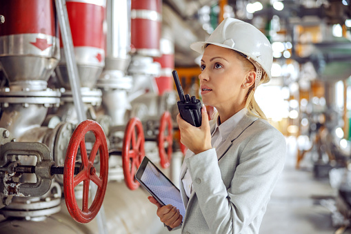 Middle aged experienced hardworking female supervisor with helmet in suit holding tablet in hands and talking on walkie talkie with employee while standing in heating plant.