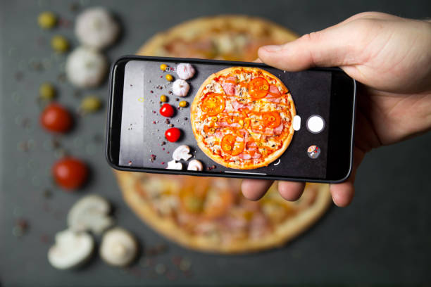 istock Using mobile phone to photograph the Pizza . Pizza with ingredients on a stone table top view. 1289720668
