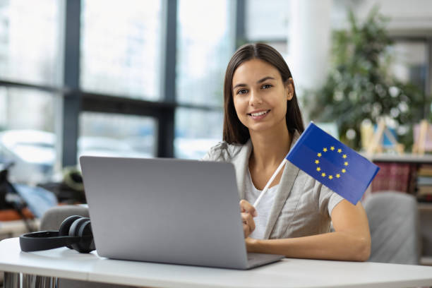 istock Beautiful female student with EU flag sitting at table with laptop and smiling while looking at camera EU study or student exchange program concept 1289722964