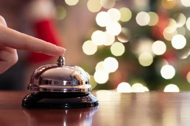 Christmas travel. Hand of guest ringing bell on reception desk of guesthouse and color shining garland on christmas tree on background. Hotel, restaurant.