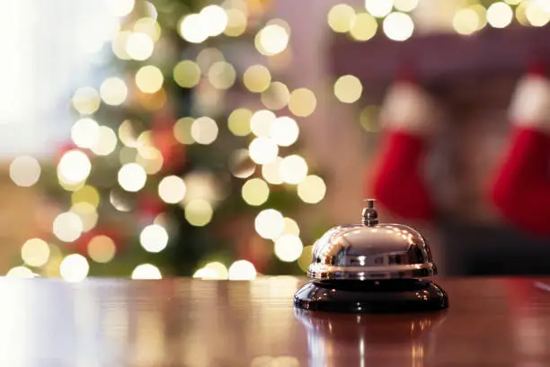 Photo of Christmas travel. Silver vintage bell on reception desk of guesthouse  and color shining garland on christmas tree on background. Hotel, restaurant.