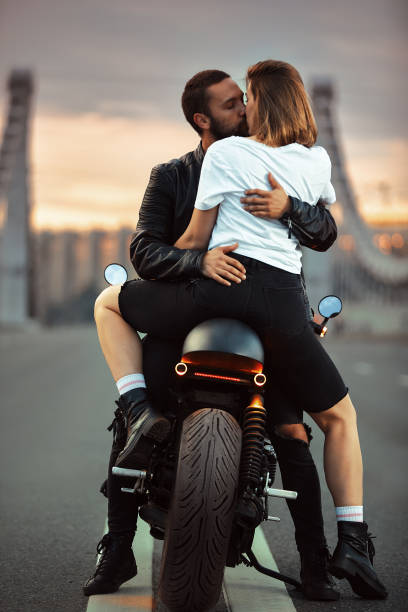 giovane donna sexy che abbraccia un uomo carino in elegante giacca di pelle nera, seduta su una moto sportiva sul ponte della città al tramonto e baciando - heterosexual couple couple sensuality young couple foto e immagini stock